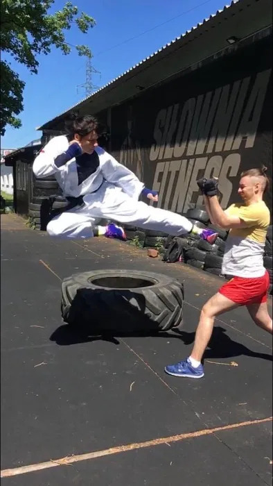 Mężczyzna w białym kimonie z czarnym pasem wykonuje wysokie kopnięcie w powietrzu, podczas sparingu z partnerem na zewnątrz siłowni z oponami w tle.