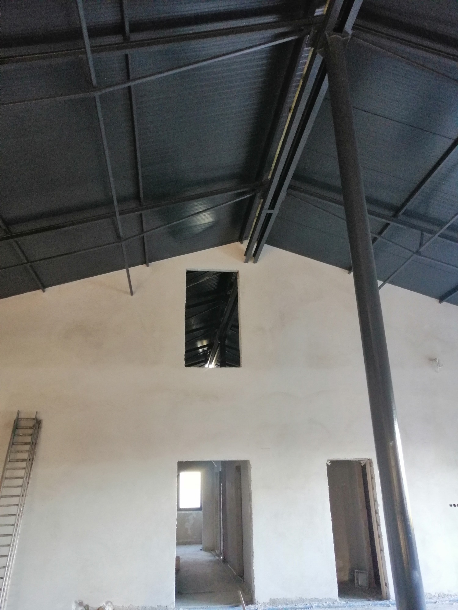 Wnętrze budynku w trakcie prac wykończeniowych: świeżo otynkowane ściany w jasnym odcieniu, widoczna metalowa konstrukcja dachu oraz drabina oparta o ścianę, trzy otwory drzwiowe i okienny w górnej...