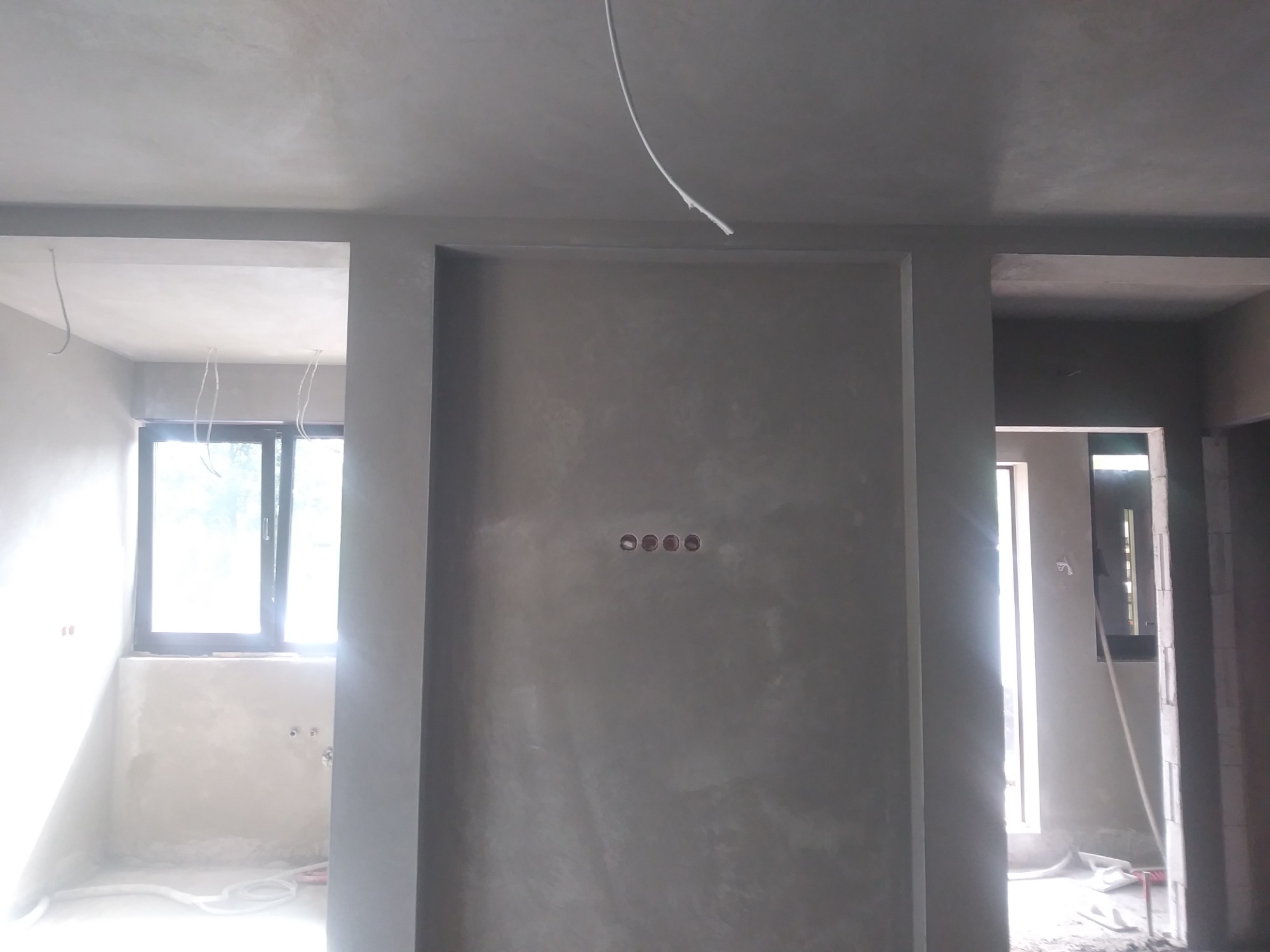 Świeżo otynkowane ściany w szarym odcieniu, widoczne otwory pod gniazdka elektryczne i fragmenty okien w jasnych pomieszczeniach obok, przewody zwisające z sufitu.