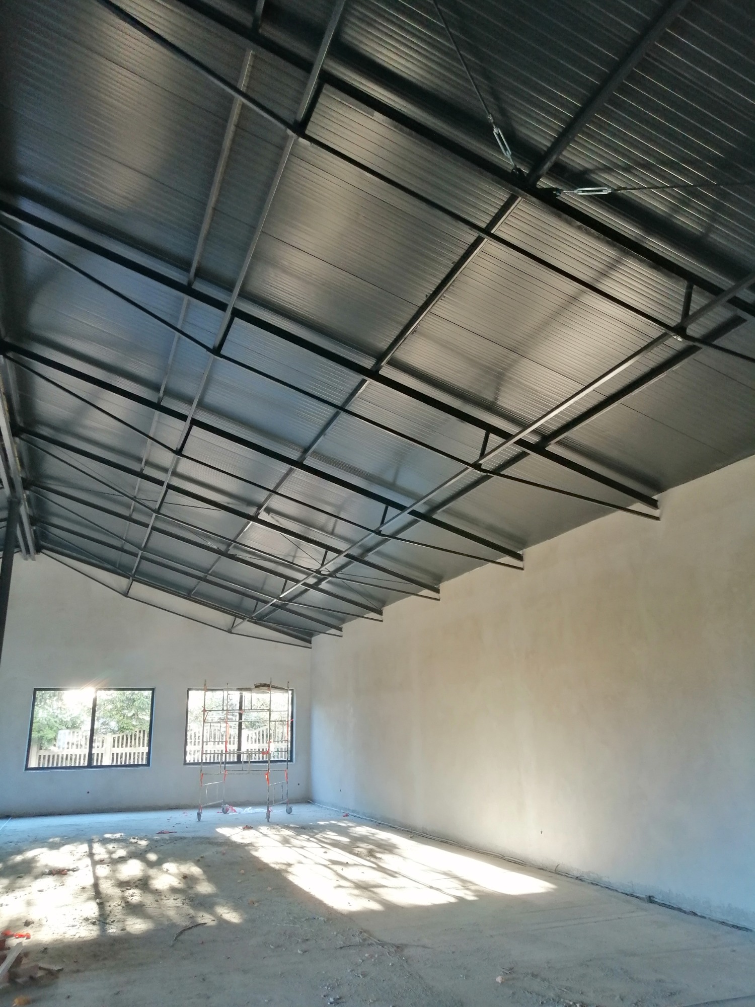 Wnętrze budynku w stanie surowym: widoczne ściany po nałożeniu wstępnej warstwy, metalowa konstrukcja dachu z blachy trapezowej, oraz rusztowanie pod oknem.