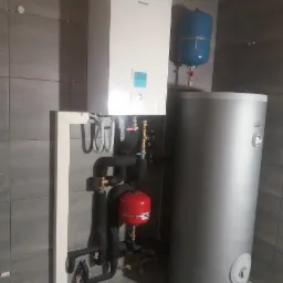 Biała pompa ciepła Panasonic z podłączonymi rurami w izolacji, czerwonym zbiornikiem wyrównawczym i szarym zasobnikiem ciepłej wody, zamontowana w pomieszczeniu wyłożonym szarymi płytkami.