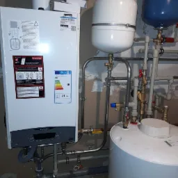 Biały piec gazowy Ariston z podłączonymi rurami i zbiornikami wyrównawczymi w pomieszczeniu z surowymi ścianami.