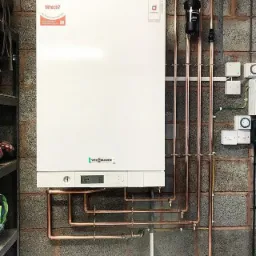 Biały piec grzewczy Viessmann zamontowany na szarej ścianie z bloków betonowych, z widocznymi miedzianymi rurami, filtrem wody i osprzętem elektrycznym. Na piecu naklejka z oceną 'Best Buy'.