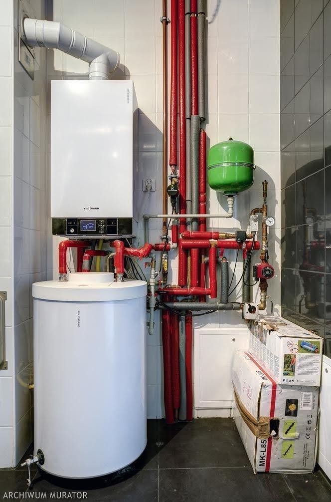 Biały, kondensacyjny kocioł gazowy Viessmann z podgrzewaczem wody, zainstalowany w pomieszczeniu z białymi kafelkami, widoczne rury centralnego ogrzewania w izolacji w kolorze czerwonym i szarym...