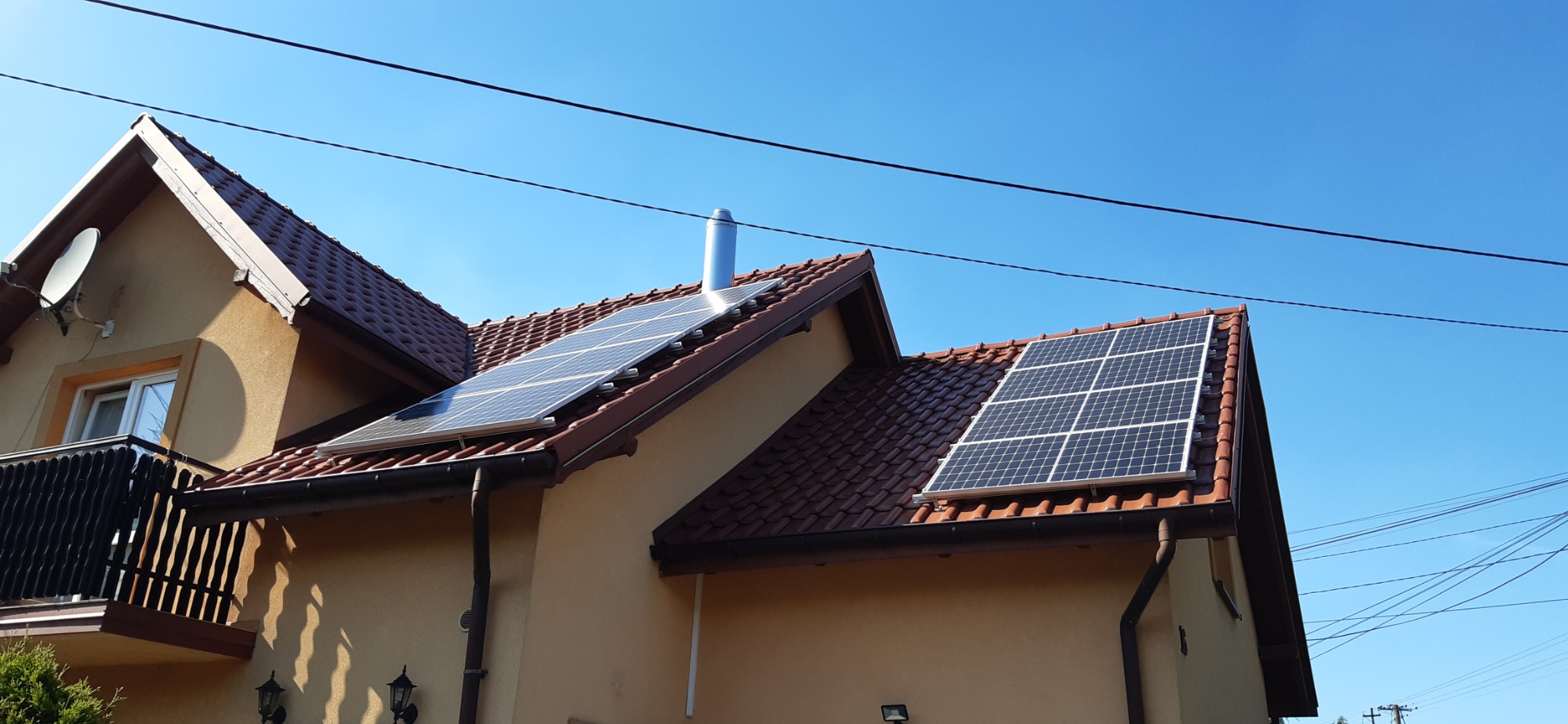 Panele fotowoltaiczne zamontowane na dachu domu jednorodzinnego z brązową dachówką, widoczne z dołu pod kątem, na tle błękitnego nieba.