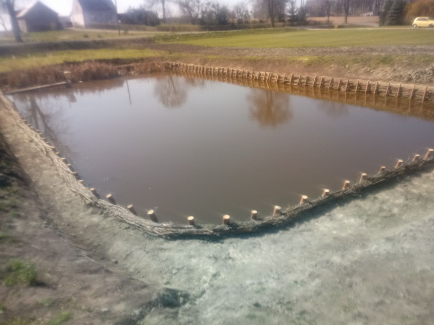 Umacnianie skarpy stawu z wykorzystaniem drewnianych pali i wikliny, widok na brzeg z odblaskami drzew na powierzchni wody.