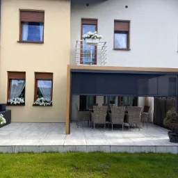 Nowoczesny taras z pergolą i meblami ogrodowymi, jasne płytki, widok na budynek z balkonem ozdobionym kwiatami, zadbany trawnik.