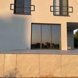 Nowoczesny taras z kamienną okładziną, zintegrowany z budynkiem, z widocznymi detalami okien i minimalistyczną architekturą w ciepłym świetle.