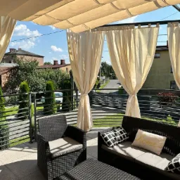 Zadaszony taras z metalową konstrukcją, beżowymi zasłonami, meblami ogrodowymi z rattanu i widokiem na okolicę.