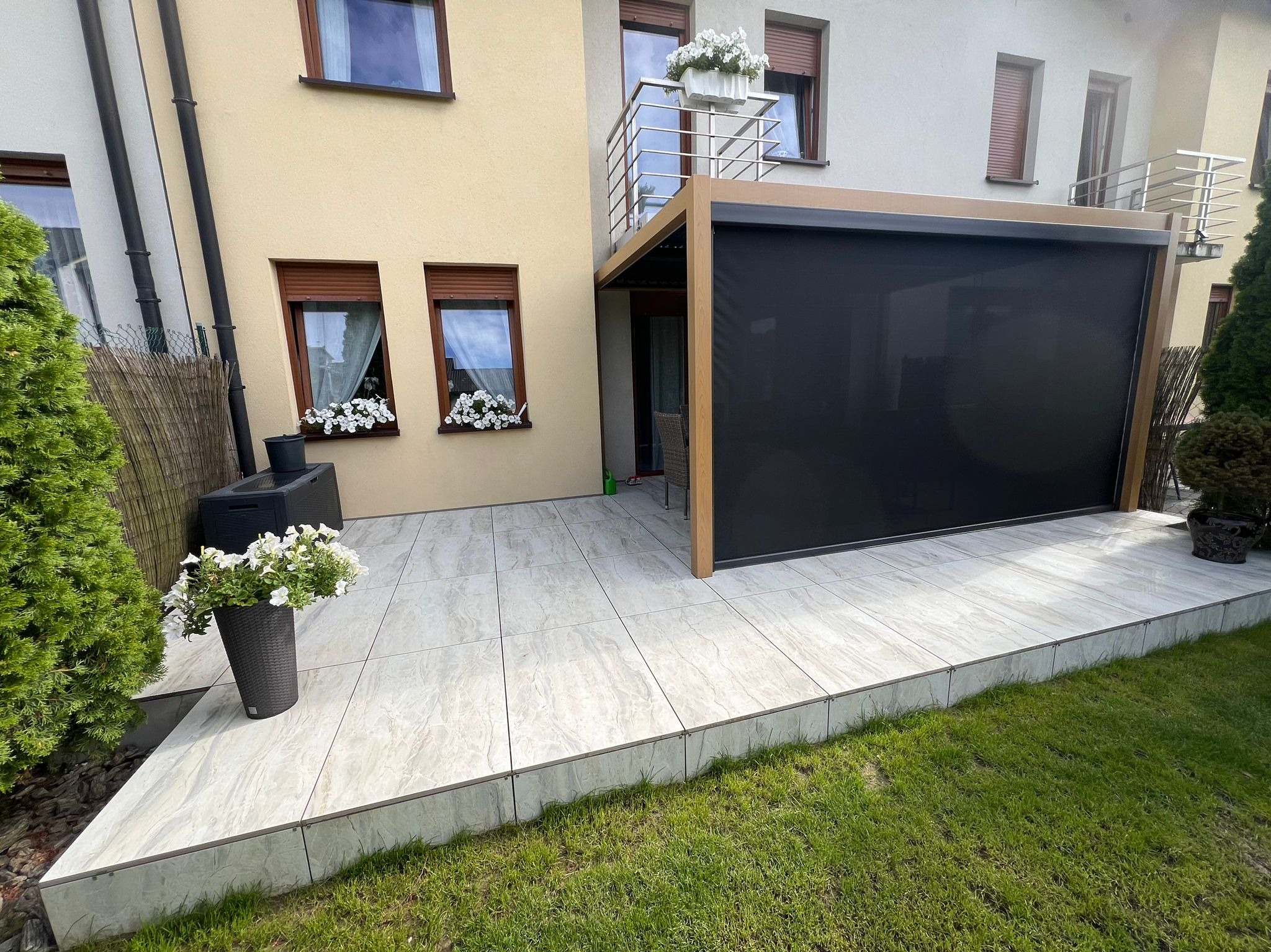 Nowoczesny taras z jasnych płyt, z pergolą z czarną roletą, balkonem powyżej i kwiatami w oknach. Trawa wokół tarasu.