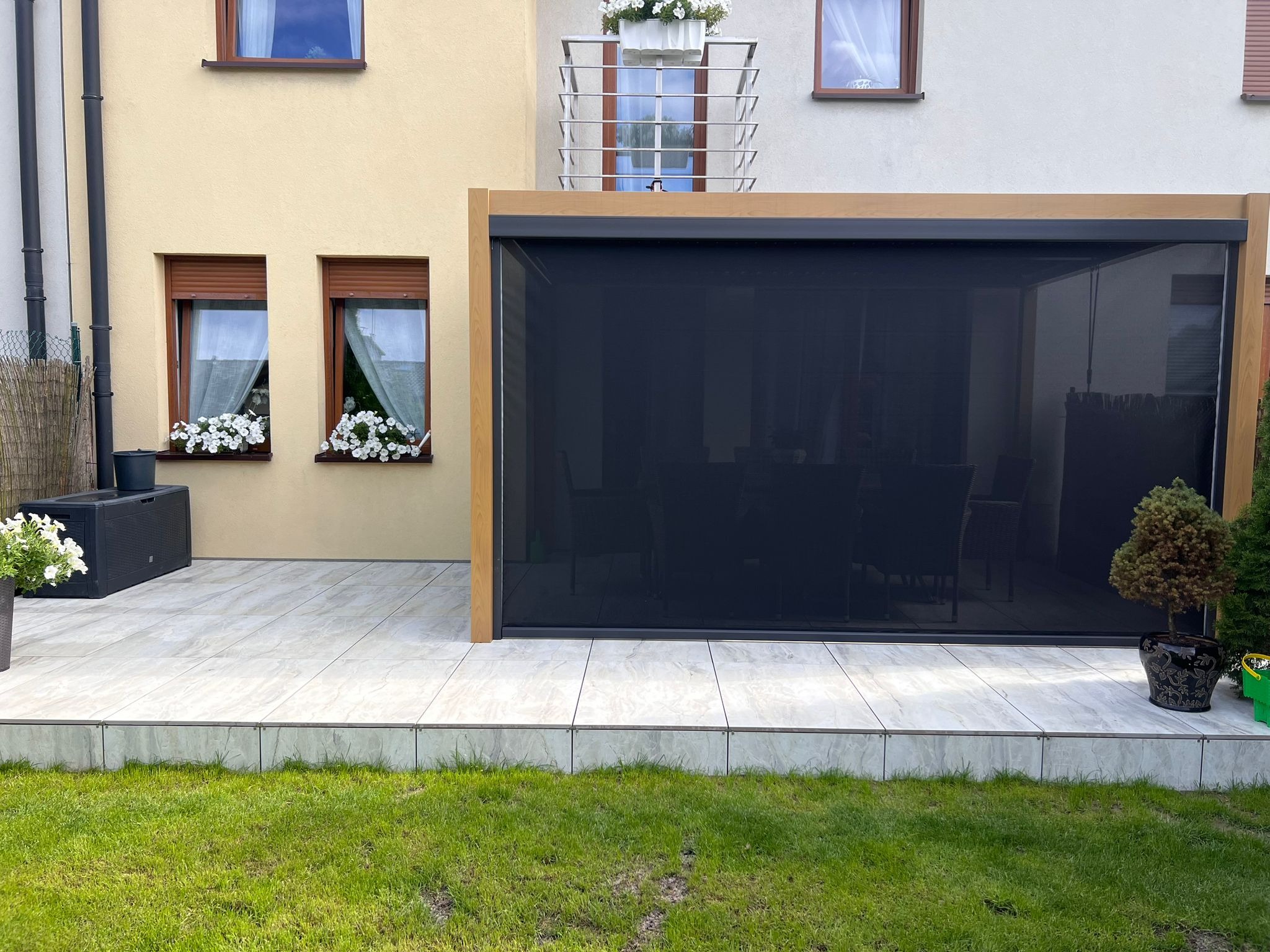 Nowoczesna pergola z zasłoną na tarasie z jasnych płytek. Widoczne kwiaty w oknach i donicach, oraz trawnik. Ujęcie z poziomu trawy.