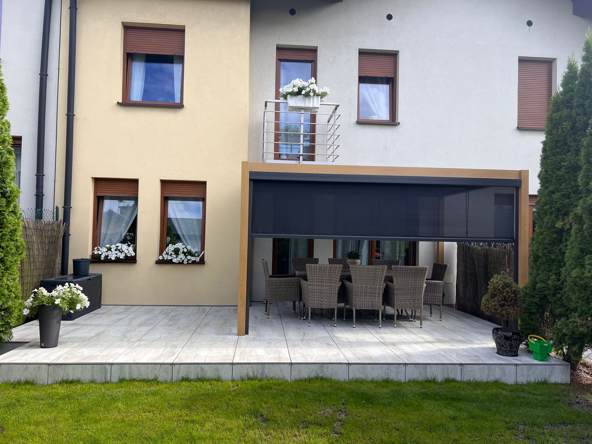 Nowoczesny taras z pergolą i meblami ogrodowymi, jasne płytki, widok na budynek z balkonem ozdobionym kwiatami, zadbany trawnik.