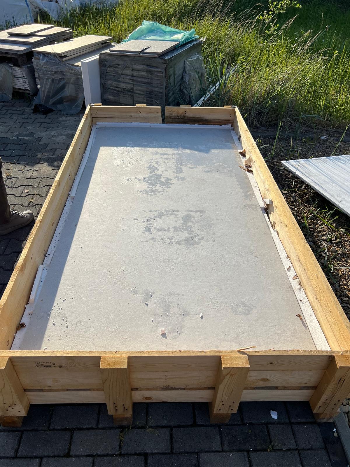 Płyta betonowa w drewnianej skrzyni transportowej, widoczne inne płyty w tle, trawa i kostka brukowa. Płyta ma szarą, nierówną powierzchnię.