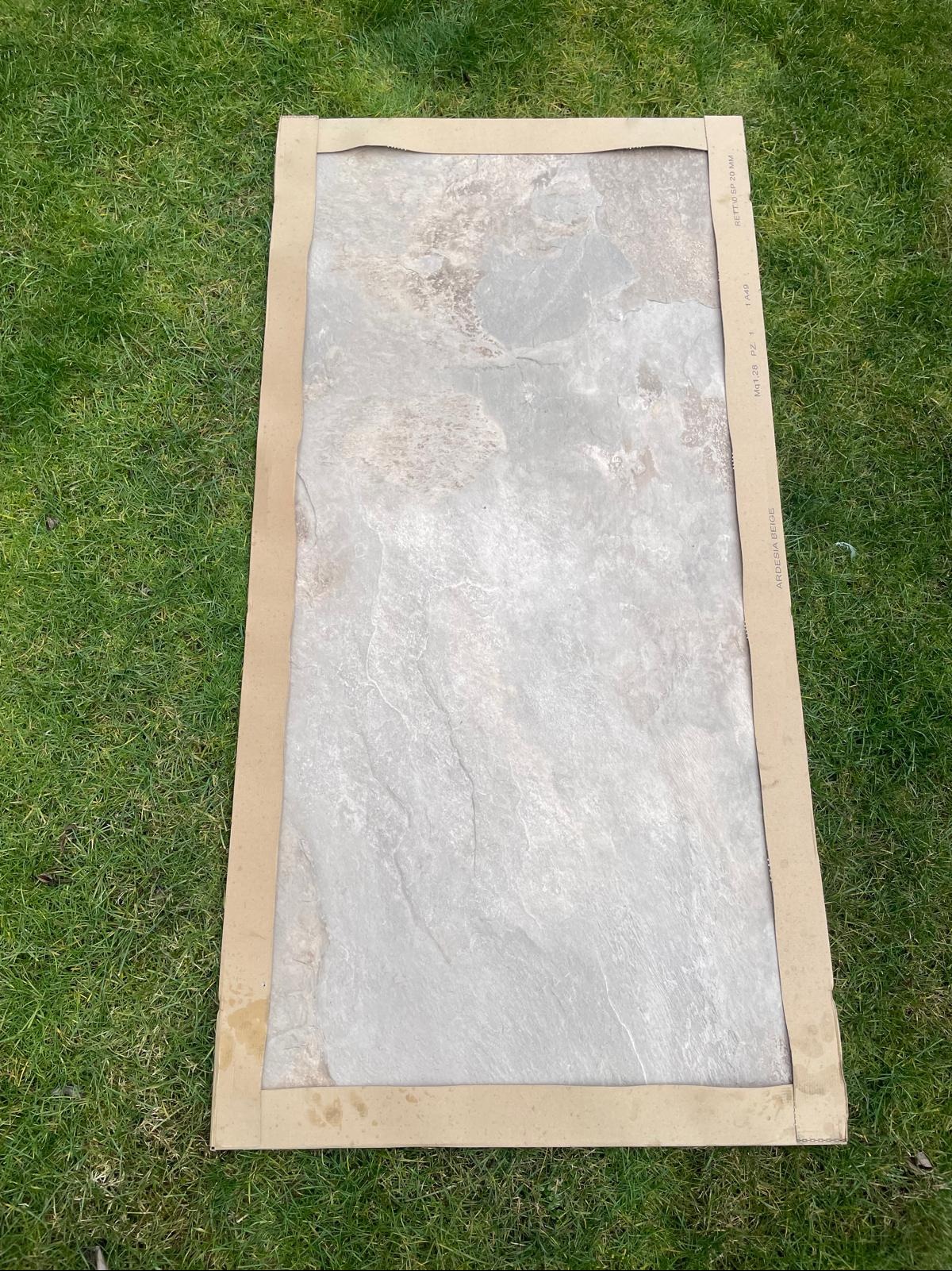 Płyta kamienna w beżowym kartonowym opakowaniu na zielonej trawie. Widoczne oznaczenia producenta na opakowaniu. Ardesia Beige.