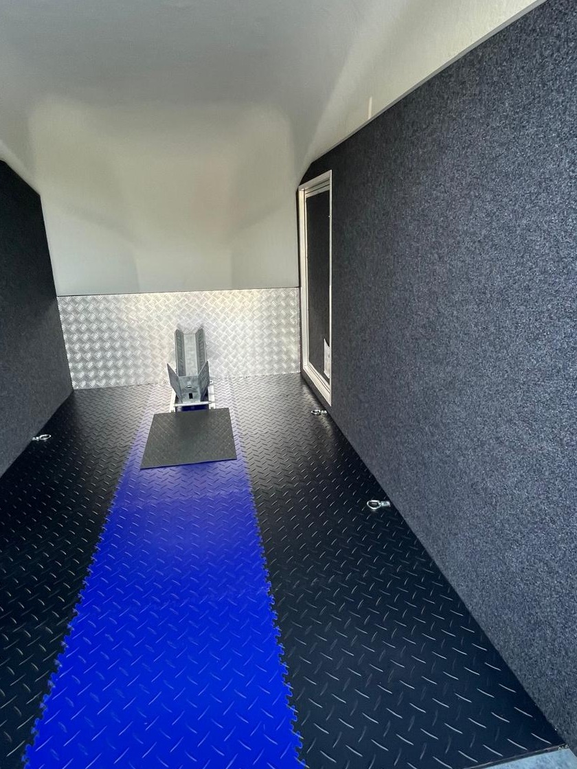 Wnętrze przyczepy z niebieską podłogą, aluminiową ścianą i uchwytem na motocykl. Ściany wyłożone szarym materiałem. Widok z perspektywy wnętrza.