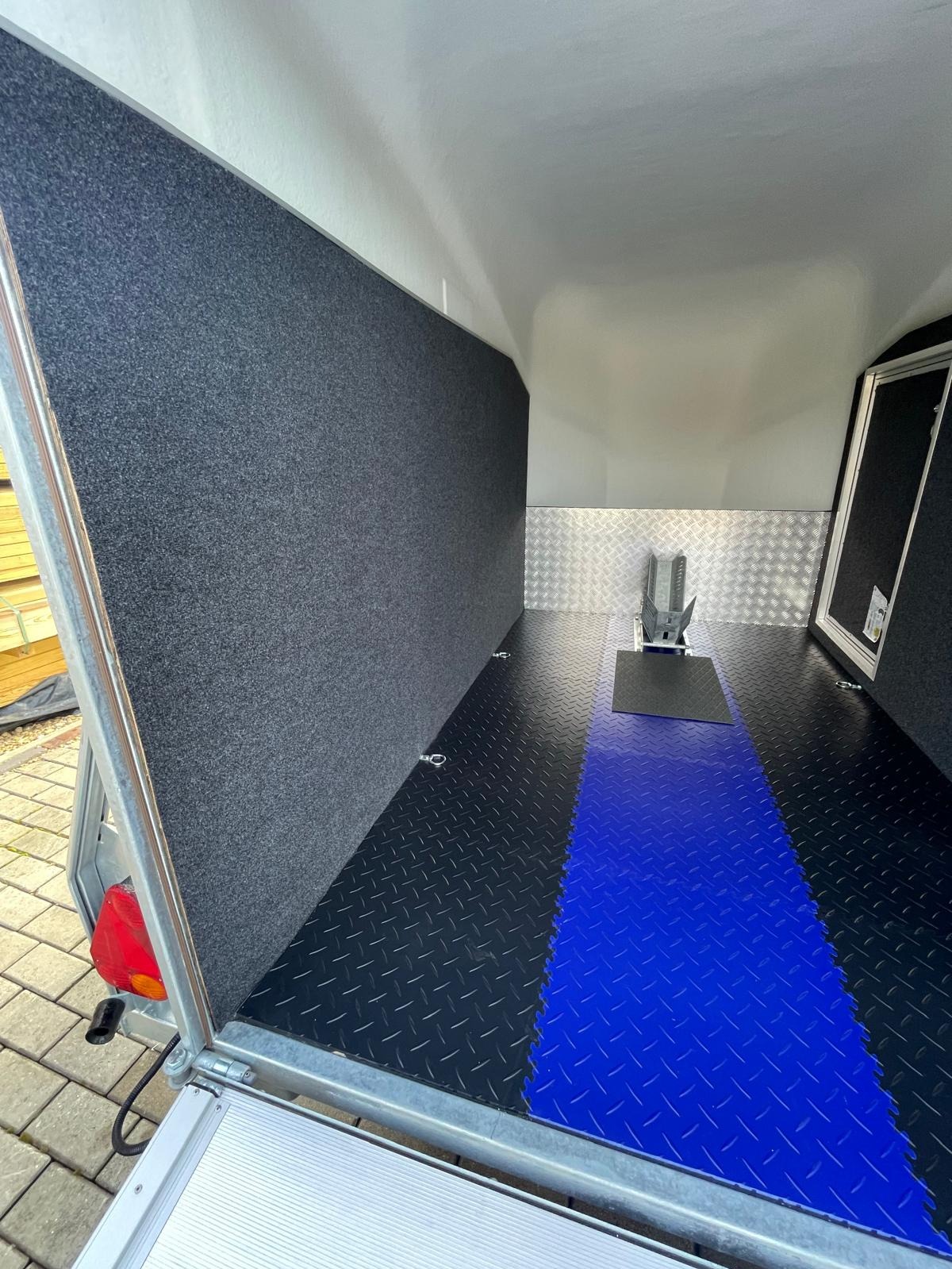 Wnętrze przyczepy z zamontowanym uchwytem na motocykl, wyłożone wykładziną i antypoślizgową podłogą w kolorze czarnym i niebieskim.