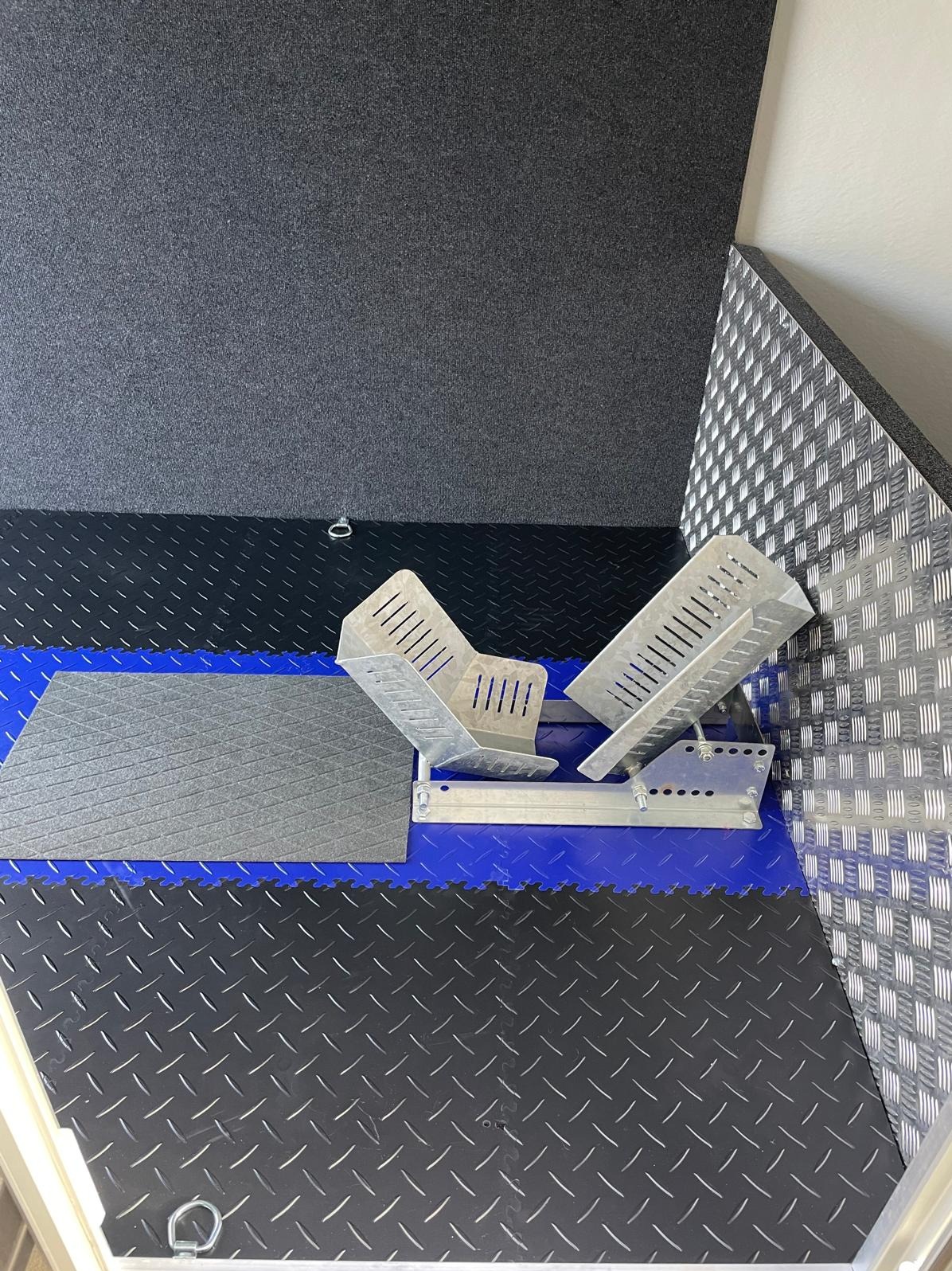 Metalowy stojak na motocykl umieszczony na podłodze z gumowych płytek w garażu. Ściany wykończone panelami o fakturze diamentowej.