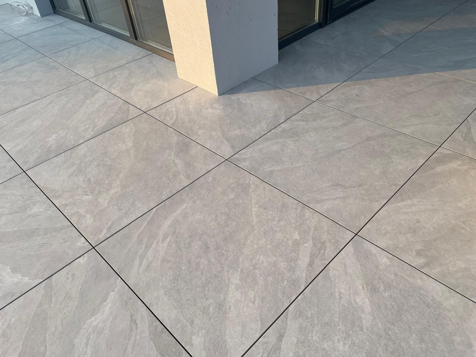 Szare płytki imitujące kamień na tarasie, ułożone równo z fugami. Widoczny fragment słupa i okna balkonowego. Poranne światło podkreśla strukturę.