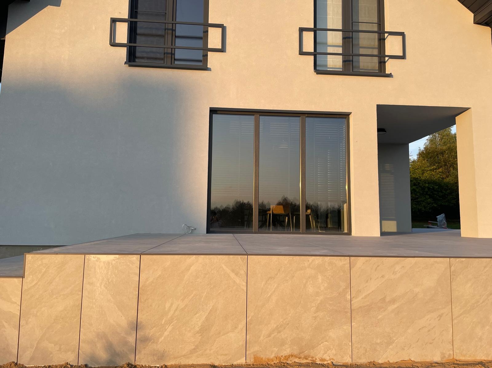Nowoczesny taras z kamienną okładziną, zintegrowany z budynkiem, z widocznymi detalami okien i minimalistyczną architekturą w ciepłym świetle.