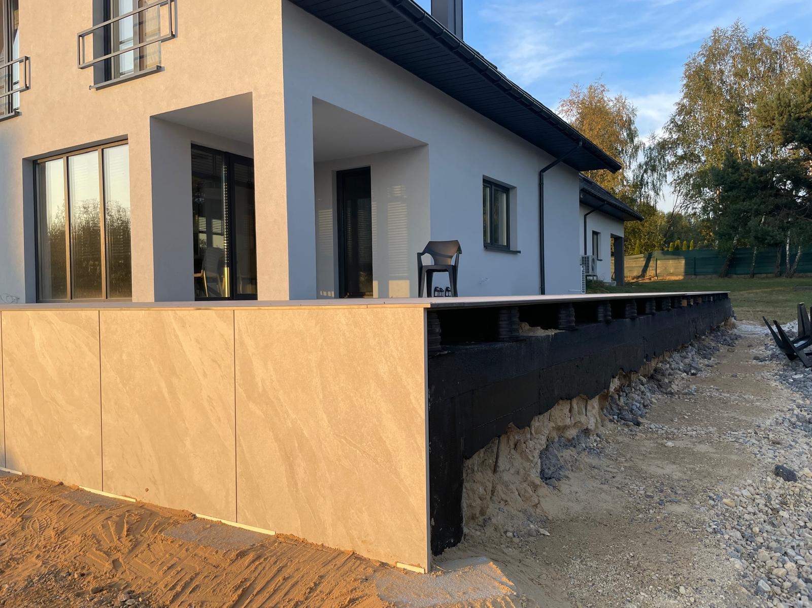 Wykończony taras z płyt kamiennych przy nowoczesnym domu. Widoczne czarne podpory tarasu i przygotowany teren pod dalsze prace. Krzesło na tarasie.