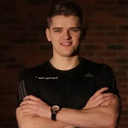 Młody mężczyzna w sportowej koszulce z logo firmy, z zegarkiem na ręku, pozuje na tle ceglanej ściany.