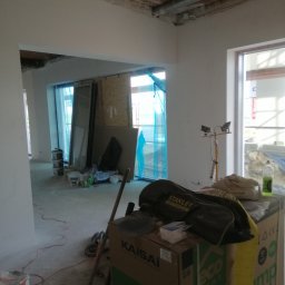 Usługi wykończeniowe - Wnętrze w trakcie remontu, widoczne świeżo otynkowane ściany, instalacja wentylacyjna pod sufitem, okna zabezpieczone folią, oraz narzędzia i materiały budowlane, w tym worki z zaprawą, kartony...