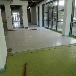 Przygotowanie podłoża pod posadzkę w biurowcu: zielona mata izolacyjna na podłodze, częściowo pokryta płytkami posadzkowymi, poziomice leżące na płytkach, widoczne słupy i okna.