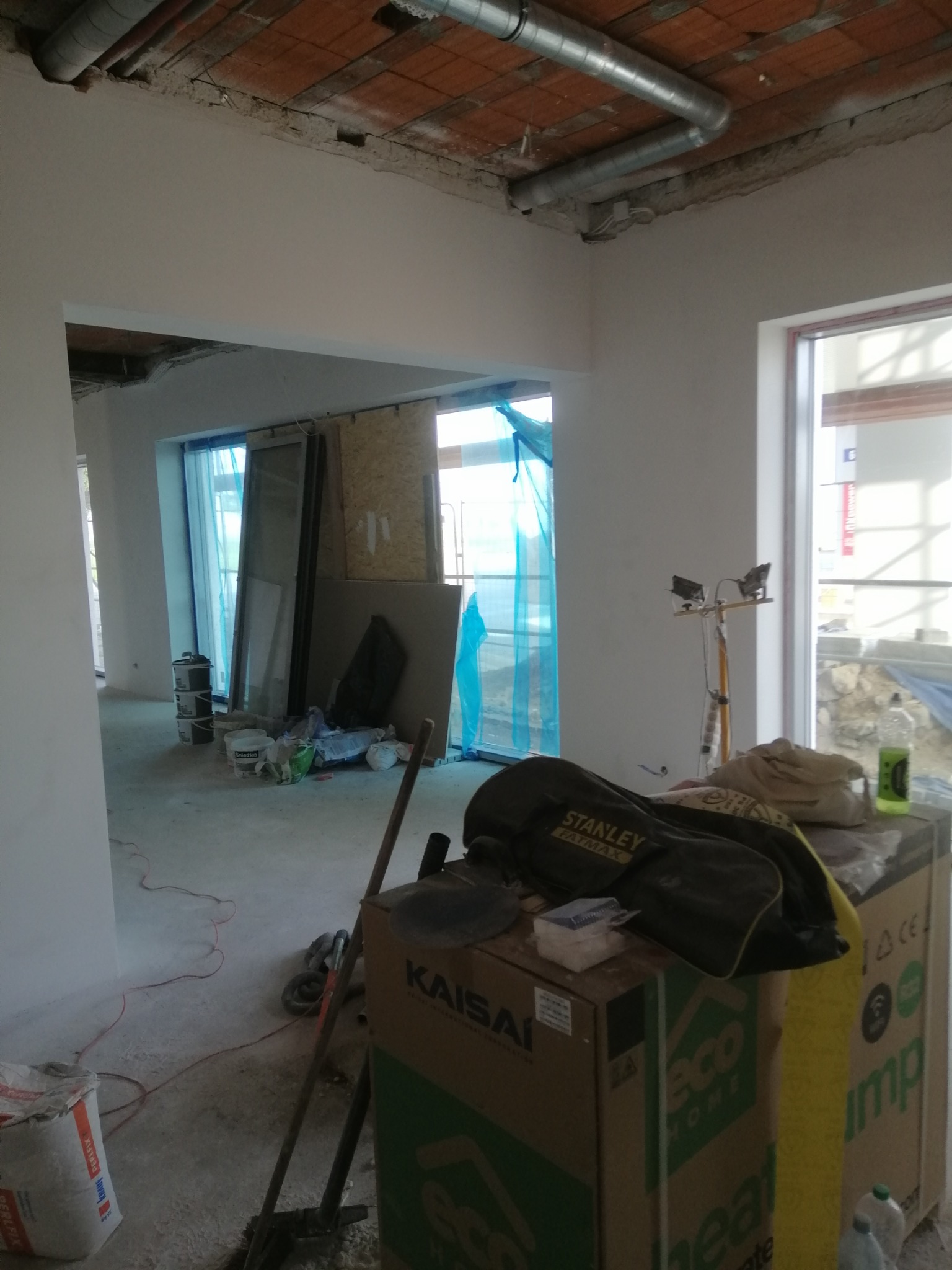 Wnętrze w trakcie remontu, widoczne świeżo otynkowane ściany, instalacja wentylacyjna pod sufitem, okna zabezpieczone folią, oraz narzędzia i materiały budowlane, w tym worki z zaprawą, kartony...