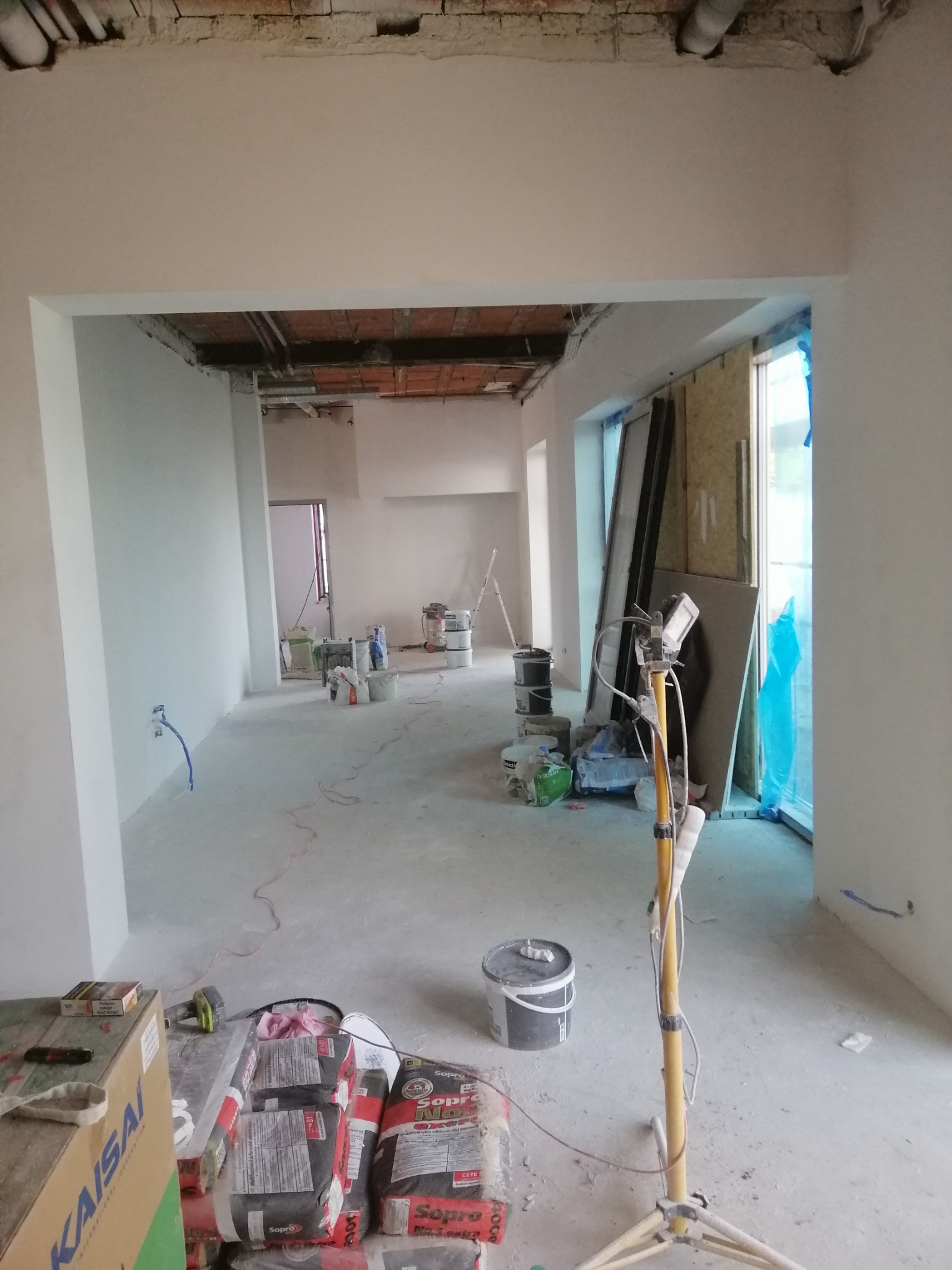 Wnętrze w trakcie prac wykończeniowych: świeżo pomalowane ściany, widoczne worki z zaprawą, wiaderka z farbą, reflektor na statywie i niezabezpieczone okno folią.