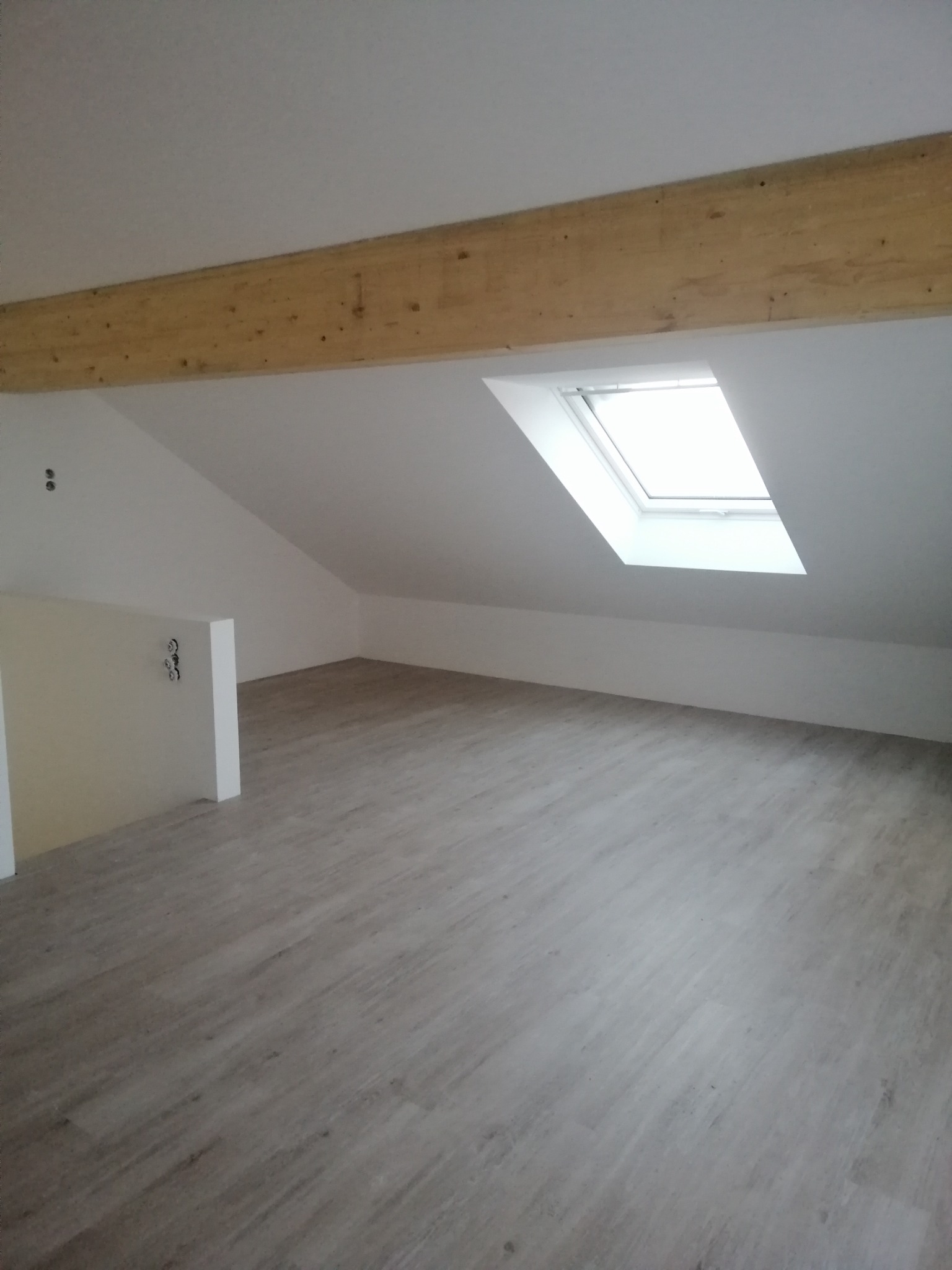 Poddasze po remoncie z jasnymi panelami podłogowymi, drewnianą belką stropową i oknem dachowym w skosie.