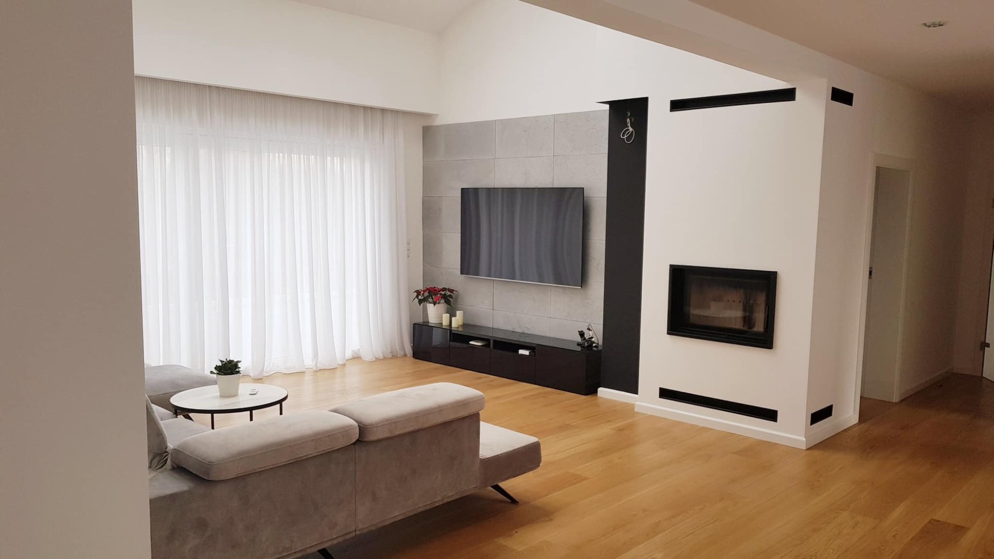 Jasny salon z drewnianą podłogą, szarym narożnikiem, telewizorem na ścianie wyłożonej szarymi płytami, kominkiem obudowanym białą ścianą z czarnymi akcentami i białymi, zwiewnymi zasłonami.
