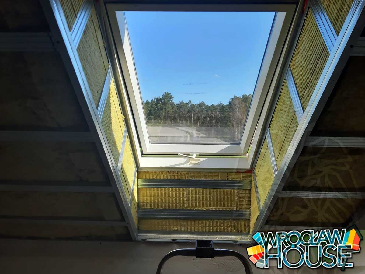 Ocieplenie poddasza wełną mineralną wokół okna dachowego, widoczne metalowe profile konstrukcyjne i fragment nieotynkowanej ściany. Na zewnątrz błękitne niebo i zieleń drzew.