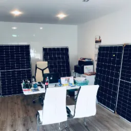 Wnętrze biura z ustawionymi pionowo panelami słonecznymi, biurkiem z laptopem i krzesłami, drewniana podłoga.