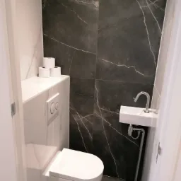 Nowoczesna, mała łazienka z czarnymi płytkami imitującymi marmur, wiszącą toaletą, umywalką narożną i zapasem papieru toaletowego na półce.