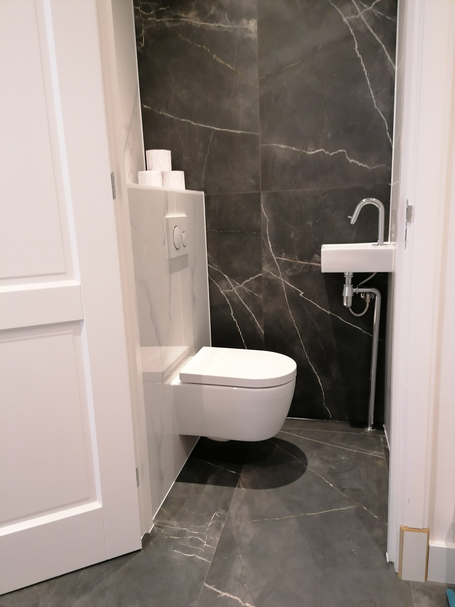 Nowoczesna łazienka z wiszącą toaletą, umywalką z chromowaną baterią i czarnymi płytkami imitującymi marmur z białymi żyłami, uzupełniona o białe elementy zabudowy i drzwi.