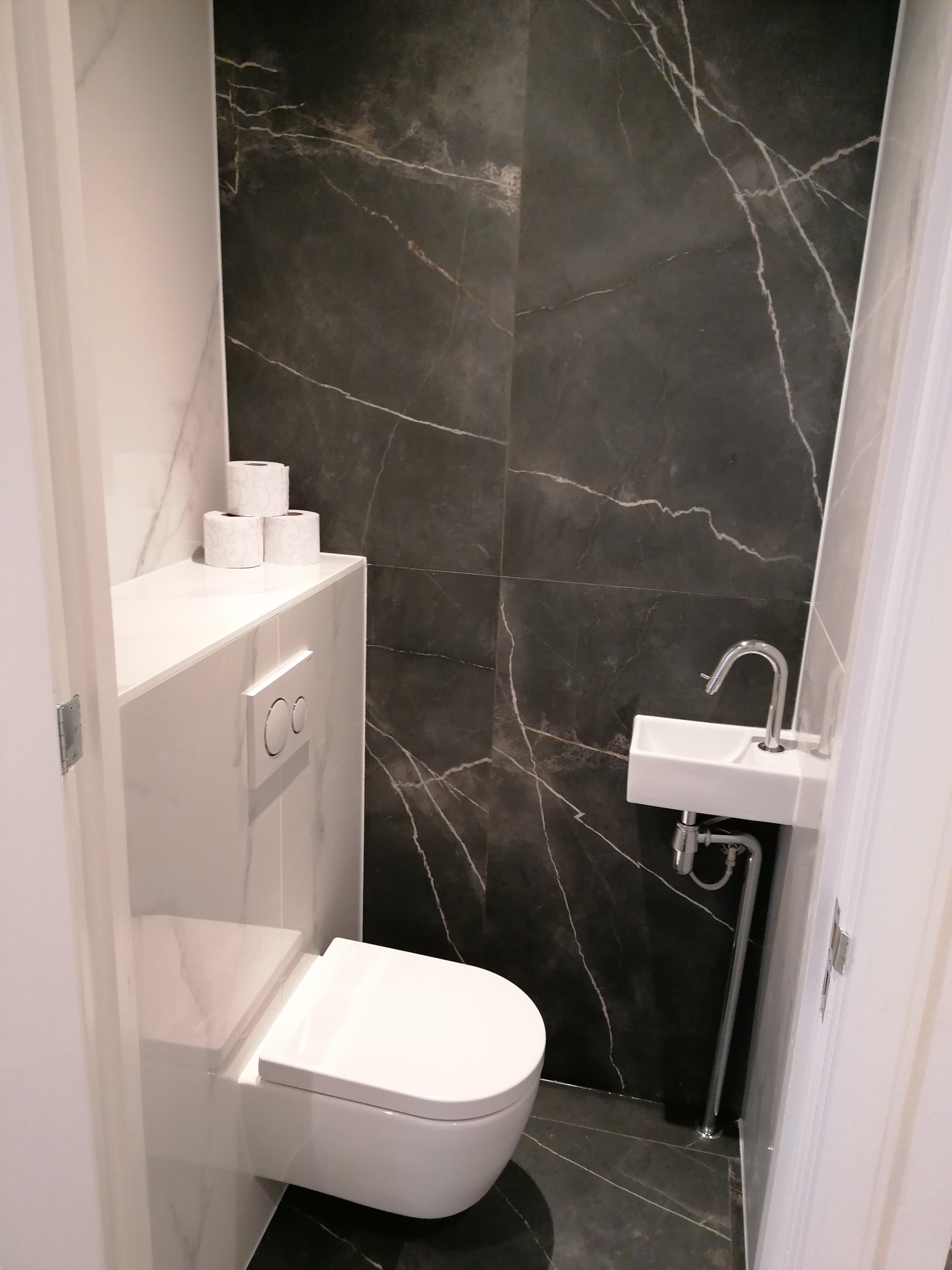 Nowoczesna, mała łazienka z czarnymi płytkami imitującymi marmur, wiszącą toaletą, umywalką narożną i zapasem papieru toaletowego na półce.