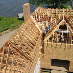 Andrzej Czarkowski - Budowa domu w technologii szkieletowej, widok z góry na konstrukcję dachu z drewnianych belek, pracownicy w niebieskich koszulkach montują elementy, w tle brzeg jeziora i zieleń.