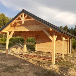 Konstrukcja drewnianej altany z czarnym dachem w trakcie budowy, widoczne elementy dekoracyjne na szczycie, stos desek i drabina oparta o pryzmę piasku.