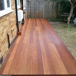 Andrzej Czarkowski - Nowo wybudowany taras z desek w odcieniach brązu, przylegający do ceglanego budynku z widocznymi rurami i skrzynką, otoczony trawnikiem i drewnianym ogrodzeniem.