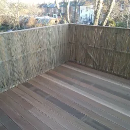 Nowoczesny taras z deski kompozytowej w odcieniach brązu i szarości, otoczony bambusową osłoną. Widok na drzewa i zabudowę miejską w tle.