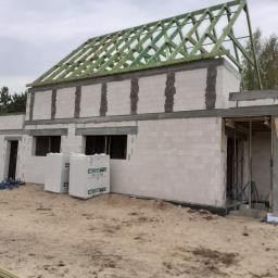 Andrzej Czarkowski - Budowa domu z białych bloczków, widoczny szkielet dachu z zielonych belek, na ziemi leżą paczki bloczków i deski.