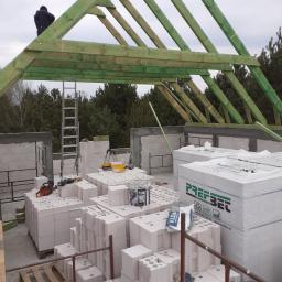 Andrzej Czarkowski - Wznoszenie konstrukcji dachu z zielonych belek na budynku z białych bloczków, widoczny pracownik na dachu, palety z materiałami budowlanymi i drabina oparta o ścianę.