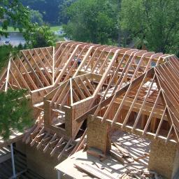 Andrzej Czarkowski - Budowa domu w technologii szkieletowej: widok z góry na drewnianą konstrukcję dachu i ścian, z elementami płyty OSB, w tle zieleń drzew i fragment jeziora.