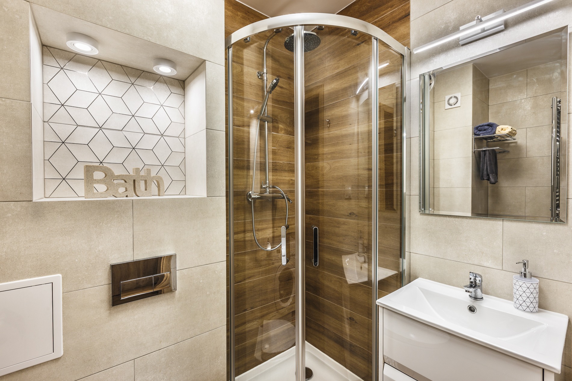 Nowoczesna łazienka z kabiną prysznicową z drewnopodobnymi panelami, umywalką z szafką, lustrem z oświetleniem LED i dekoracyjną wnęką z geometrycznymi płytkami oraz napisem 'Bath'.