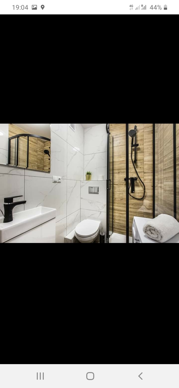 Nowoczesna łazienka w Białymstoku z białymi marmurkowymi płytkami, czarną armaturą, kabiną prysznicową z drewnopodobnymi panelami i minimalistyczną umywalką.