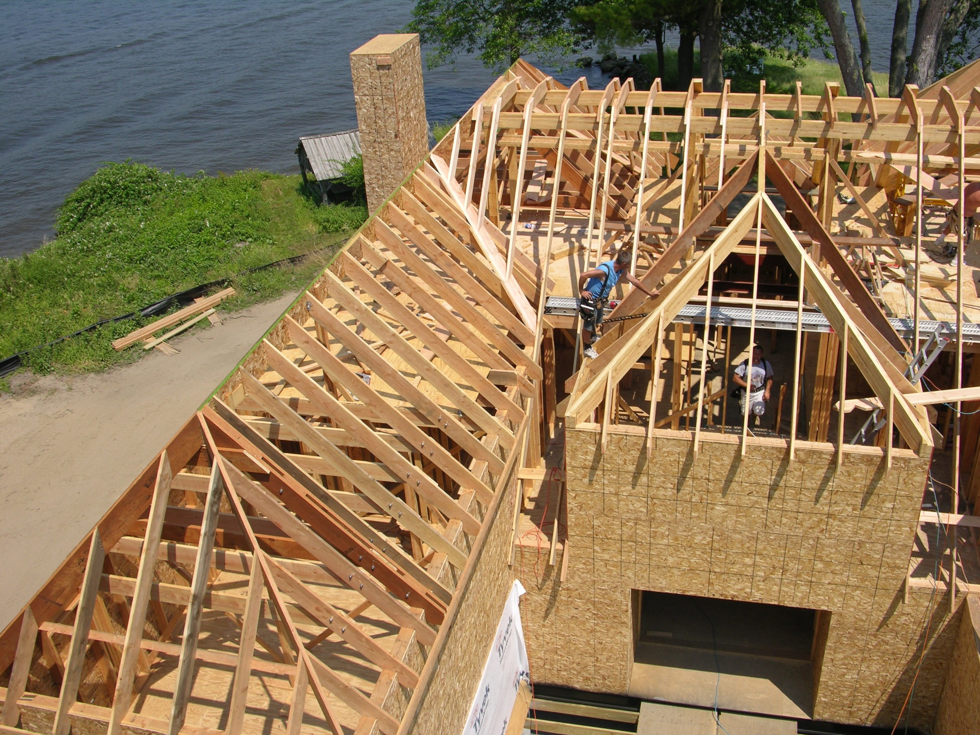 Budowa domu w technologii szkieletowej, widok z góry na konstrukcję dachu z drewnianych belek, pracownicy w niebieskich koszulkach montują elementy, w tle brzeg jeziora i zieleń.