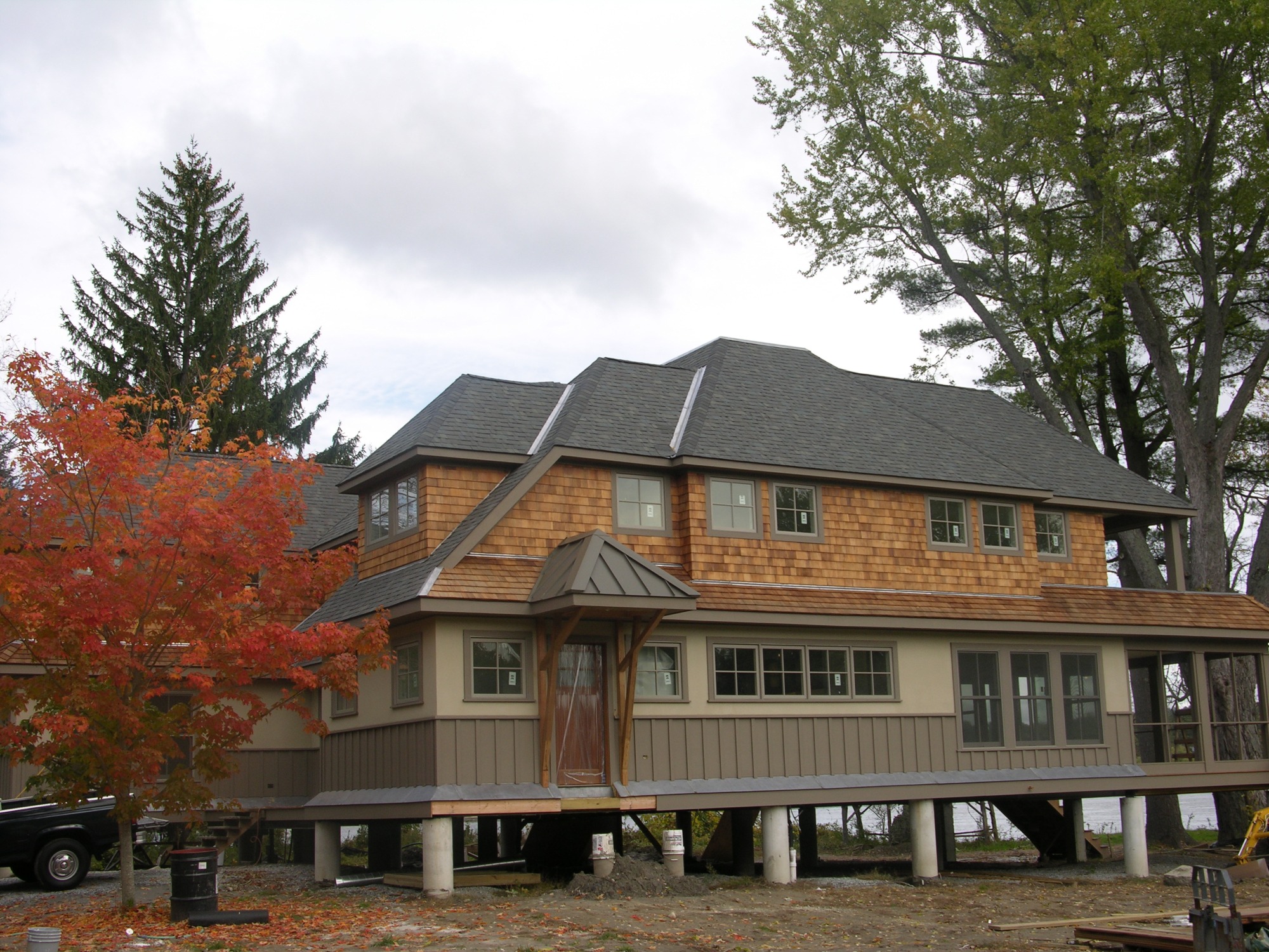 Dwupiętrowy dom na betonowych podporach, z drewnianą elewacją i szarym dachem, otoczony drzewami o jesiennych barwach, częściowo wykończony.
