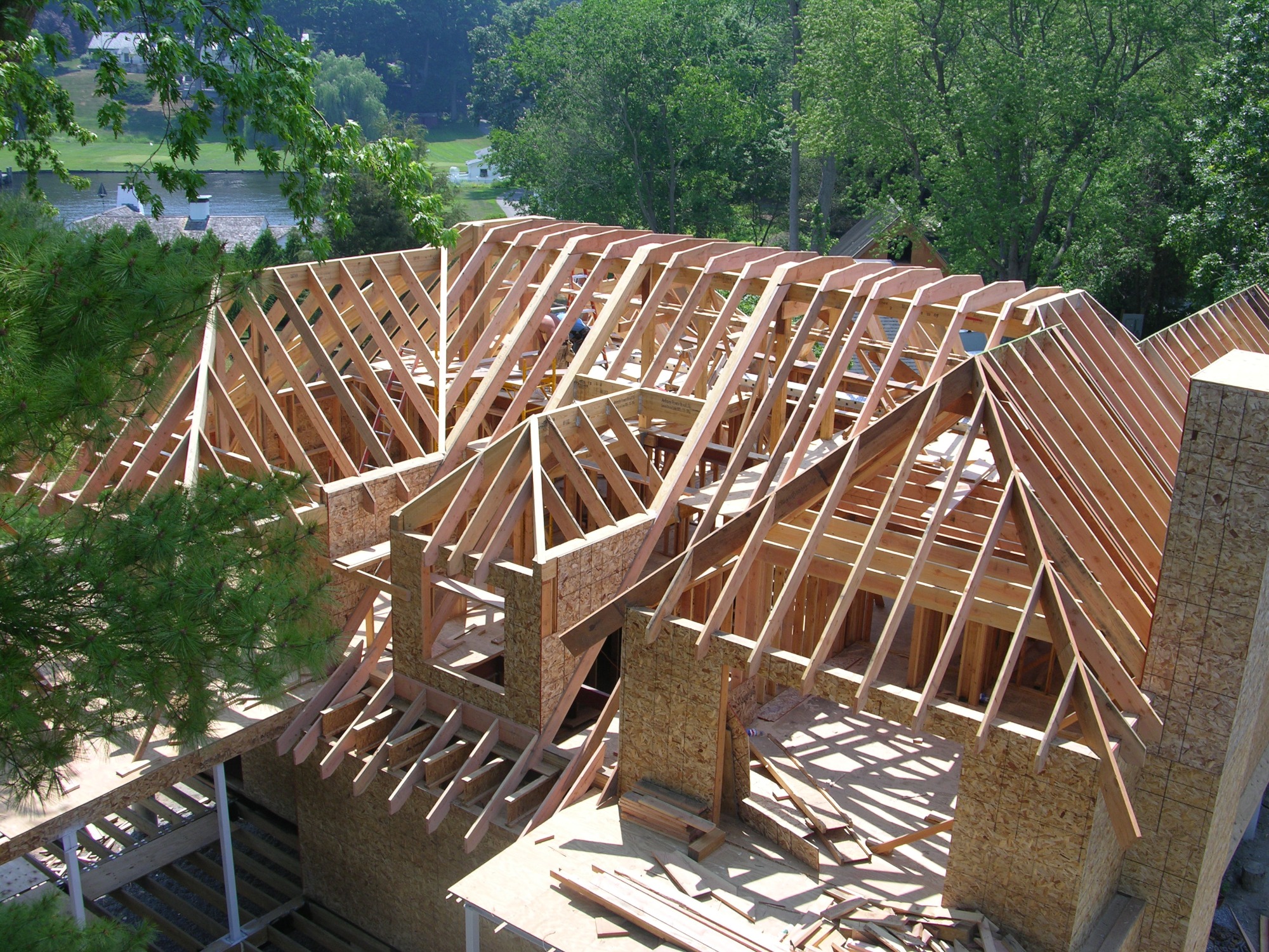 Budowa domu w technologii szkieletowej: widok z góry na drewnianą konstrukcję dachu i ścian, z elementami płyty OSB, w tle zieleń drzew i fragment jeziora.