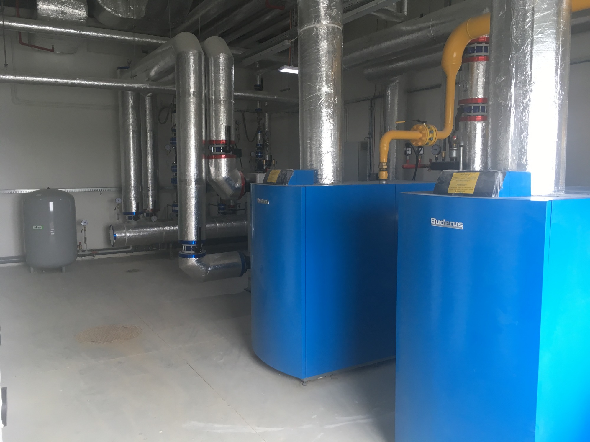 Kotłownia z dwoma niebieskimi kotłami gazowymi Buderus, srebrnymi rurami izolowanymi termicznie i szarym zbiornikiem wyrównawczym w tle, zainstalowana w obiekcie przemysłowym.
