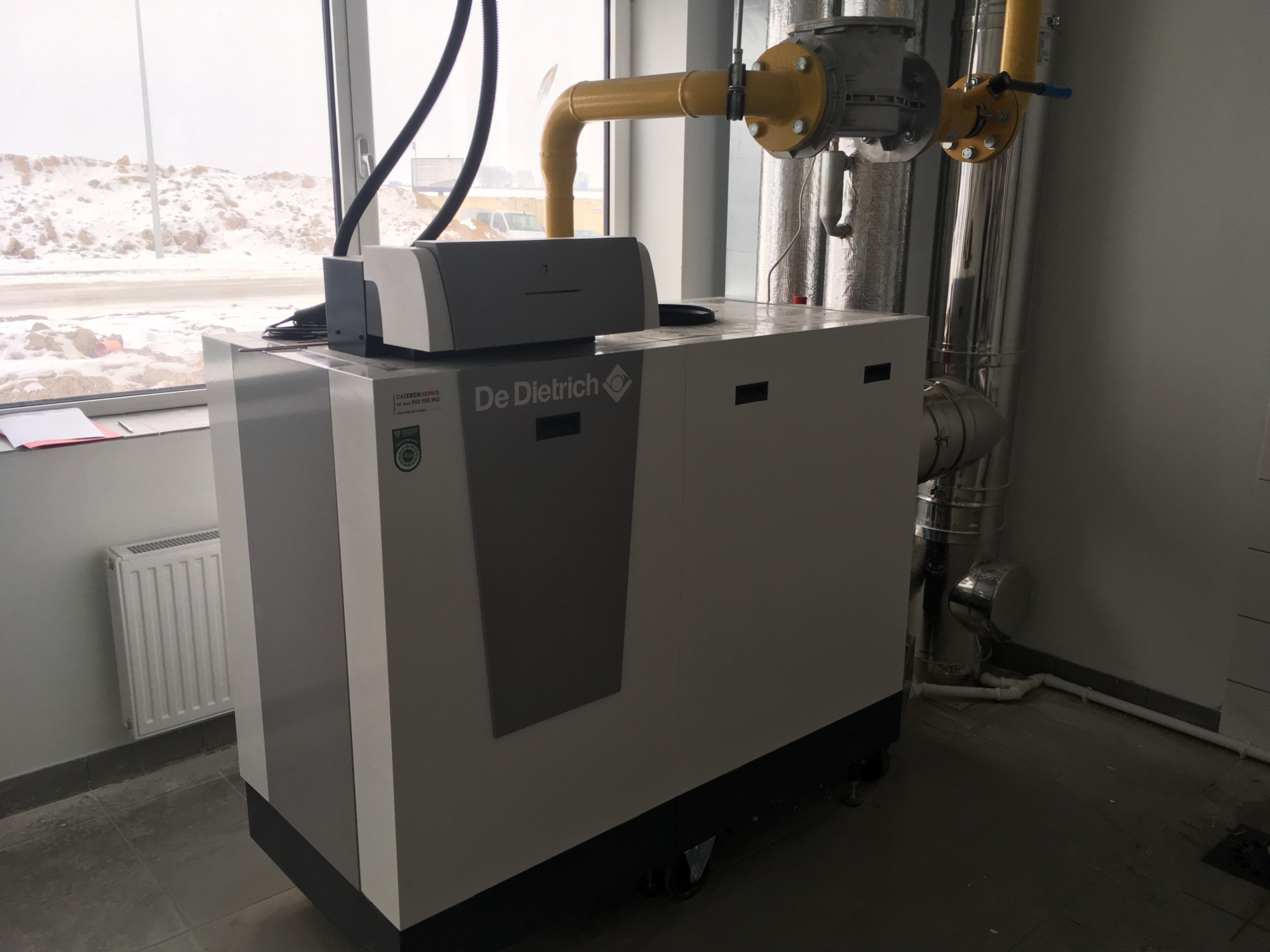 Nowoczesna kotłownia z przemysłową instalacją grzewczą De Dietrich, widoczne żółte rury gazowe, srebrne rury spalinowe, grzejnik panelowy i zaśnieżony krajobraz za oknem.